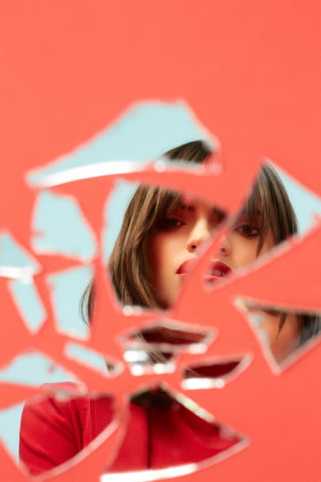 Woman's face reflected in broken mirror shards against a vibrant coral background, creating an intriguing and slightly surreal composition.