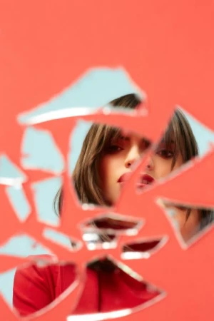 Woman's face reflected in broken mirror shards against a vibrant coral background, creating an intriguing and slightly surreal composition.