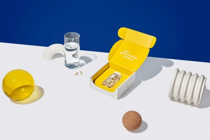 Product shot featuring an open box of dietary supplements alongside a glass of water, yellow sphere, and cylindrical decor element.