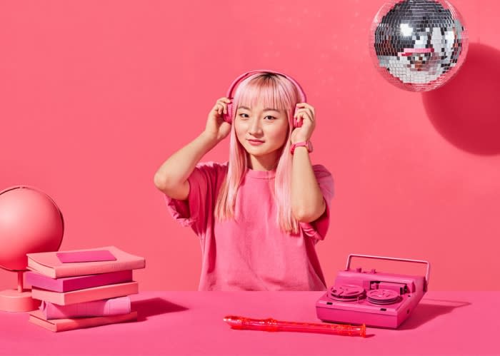 Young woman with pink hair listens to pink headphones in front of pink background with pink books, disco ball, and pink recorder.