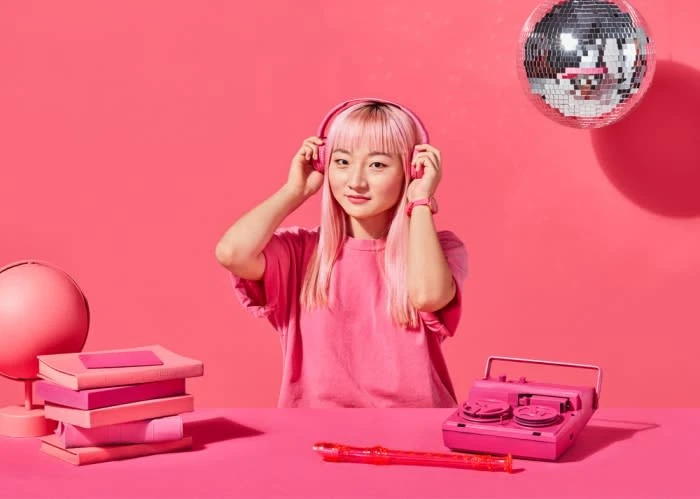 Young woman with pink hair listens to pink headphones in front of pink background with pink books, disco ball, and pink recorder.