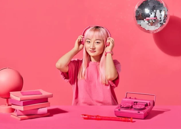Young woman with pink hair listens to pink headphones in front of pink background with pink books, disco ball, and pink recorder.