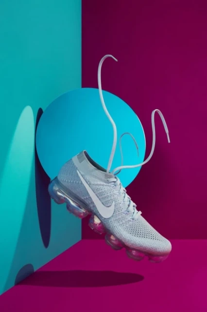 White Nike VaporMax sneaker with floating laces on a vibrant background of turquoise and magenta in a studio product shot.
