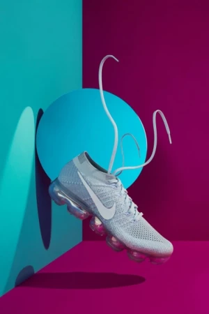 White Nike VaporMax sneaker with floating laces on a vibrant background of turquoise and magenta in a studio product shot.