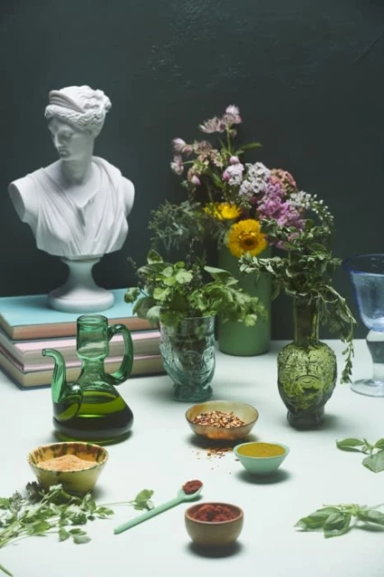 Still life arrangement with a bust of a woman, flowers, green glass vases, spices in bowls, and herbs on a white surface.