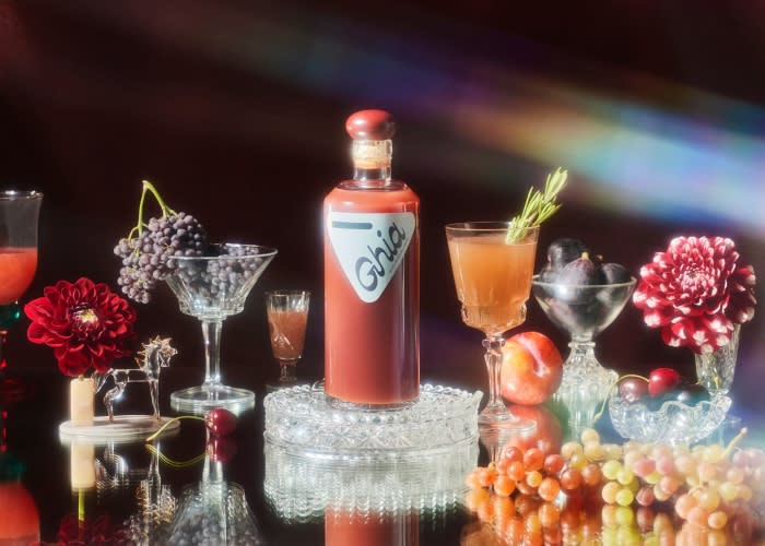 Bottle of Ghia non-alcoholic apéritif surrounded by glassware, fruit, and flowers on a reflective black surface, styled with rainbow prism lighting.