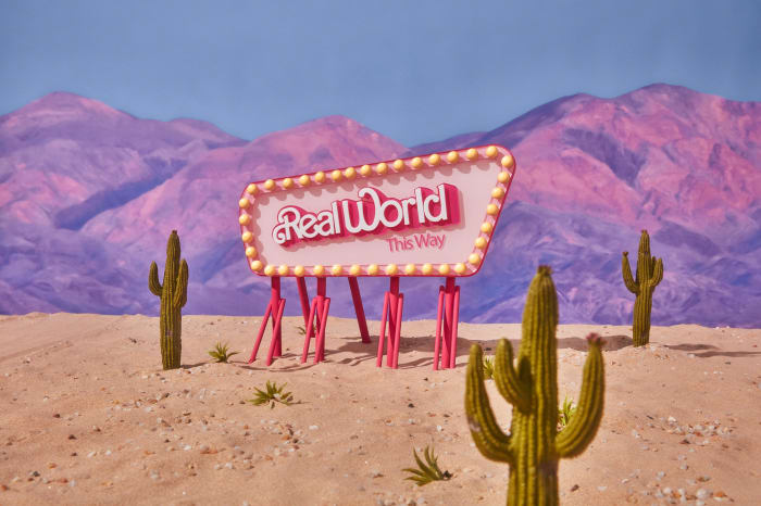 Retro style 'Real World This Way' sign in a desert landscape with cactus plants under a blue sky and purple mountains.