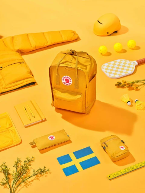 Yellow Fjallraven Kanken backpack surrounded by yellow sports equipment and school supplies on a bright yellow background in a studio setting.