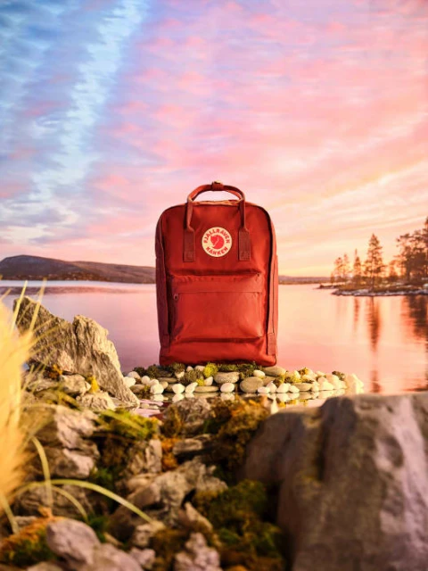 Red Fjallraven Kanken backpack displayed on a rocky surface by a tranquil lake reflecting a sunset sky with pink and blue hues.