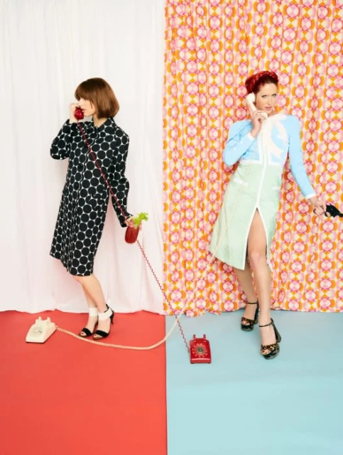Two women in retro outfits pose in a split studio space, talking on vintage phones, with a bright, vintage aesthetic and patterned backdrop.
