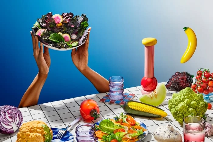 A vibrant, still life photograph of a salad in a bowl held up by two hands, surrounded by various fruits and vegetables against a blue background.