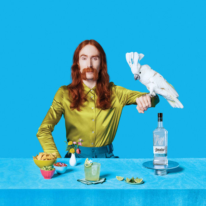 Stylish red-haired man poses with a cockatoo, accompanied by a bottle of tequila, snacks, and a cocktail on a table against a blue backdrop.