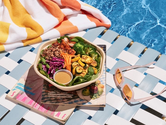 Overhead shot of a healthy salad with grilled chicken and vegetables in a bowl, with sunglasses and a striped towel by a pool.
