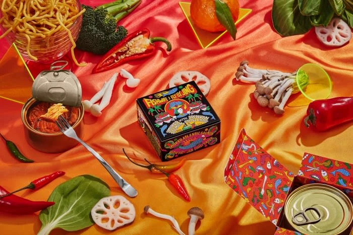 Still life food photography featuring a can of fish, noodles, vegetables, fruit, and a colorful decorative box on a textured background.