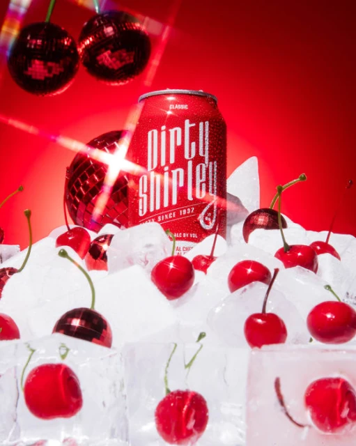 A red can of Dirty Shirley alcoholic beverage is surrounded by ice cubes and shiny cherry-shaped disco balls against a bright red backdrop.