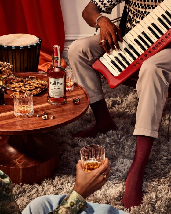 An African-American adult male plays a red keyboard on a shag rug in a room with a table, Old Forester Whiskey, and a drum.