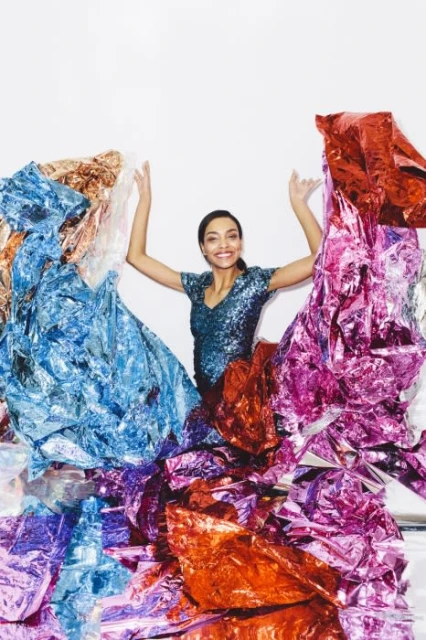 Smiling woman surrounded by shiny, colorful metallic foil in a studio setting, wearing a sparkly, v-neck top and raising her arms.