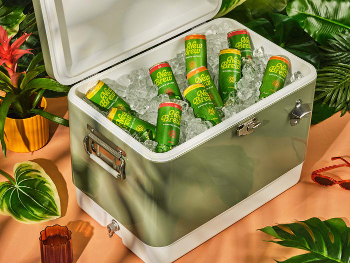 Ice-filled cooler containing multiple cans of green and gold New Brew beverage, surrounded by tropical plants and a pair of sunglasses.