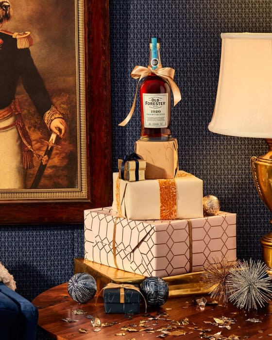 Whiskey bottle presented on stacked gifts with gold ribbon, sitting on wooden table with portrait painting in a domestic setting.