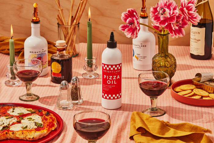 Brightland branded oils and condiments are displayed on a table with pizza, wine, flowers, and candles against a wood background.