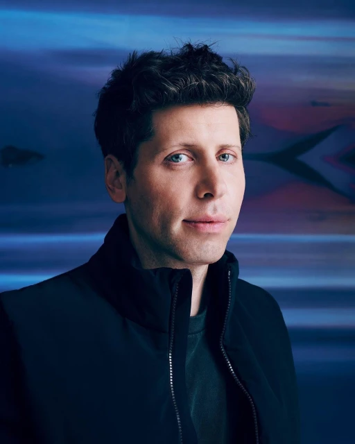 Headshot of a fair-skinned adult male with dark curly hair, wearing a black jacket against a blurred, abstract background in cool tones.
