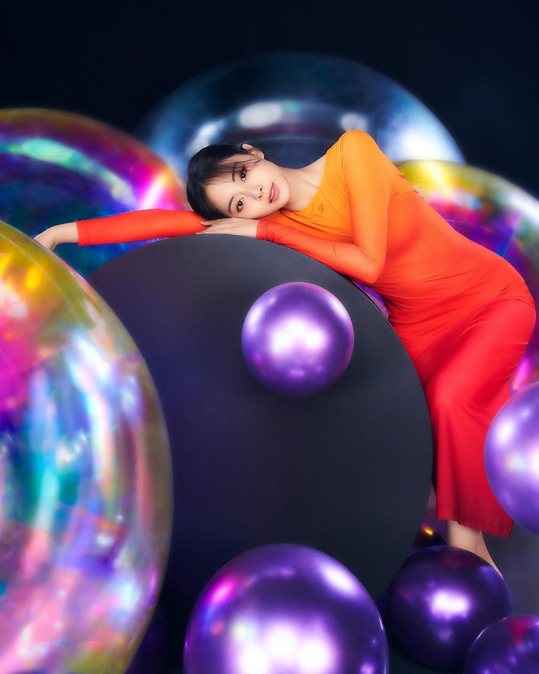 Woman in red dress poses with colorful spheres against a dark background. The spheres are iridescent and purple. The lighting is soft.
