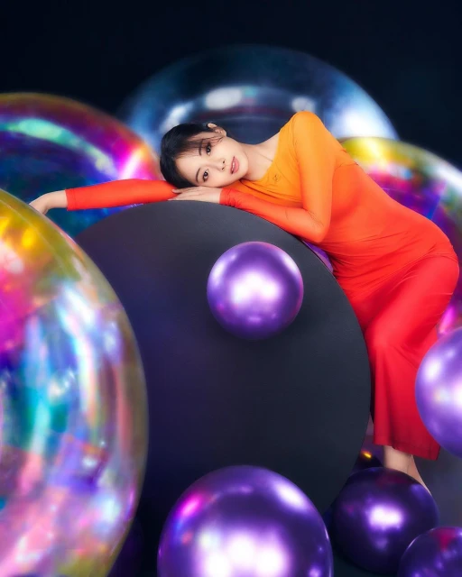 Woman in red dress poses with colorful spheres against a dark background. The spheres are iridescent and purple. The lighting is soft.