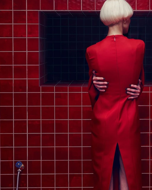 Woman with blonde hair wears a red dress with her arms crossed in a room with red tiles and a dark recessed panel.