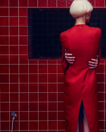 Woman with blonde hair wears a red dress with her arms crossed in a room with red tiles and a dark recessed panel.