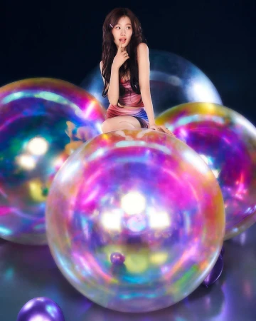 Young Asian woman poses playfully on iridescent spheres against a dark backdrop, wearing a stylish pink and blue dress.