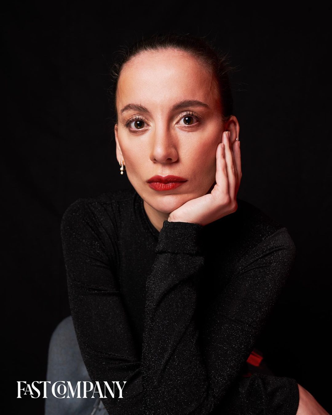 Close-up portrait of an adult woman with red lipstick and a long-sleeved black shirt against a dark background; she rests her face in her hand.