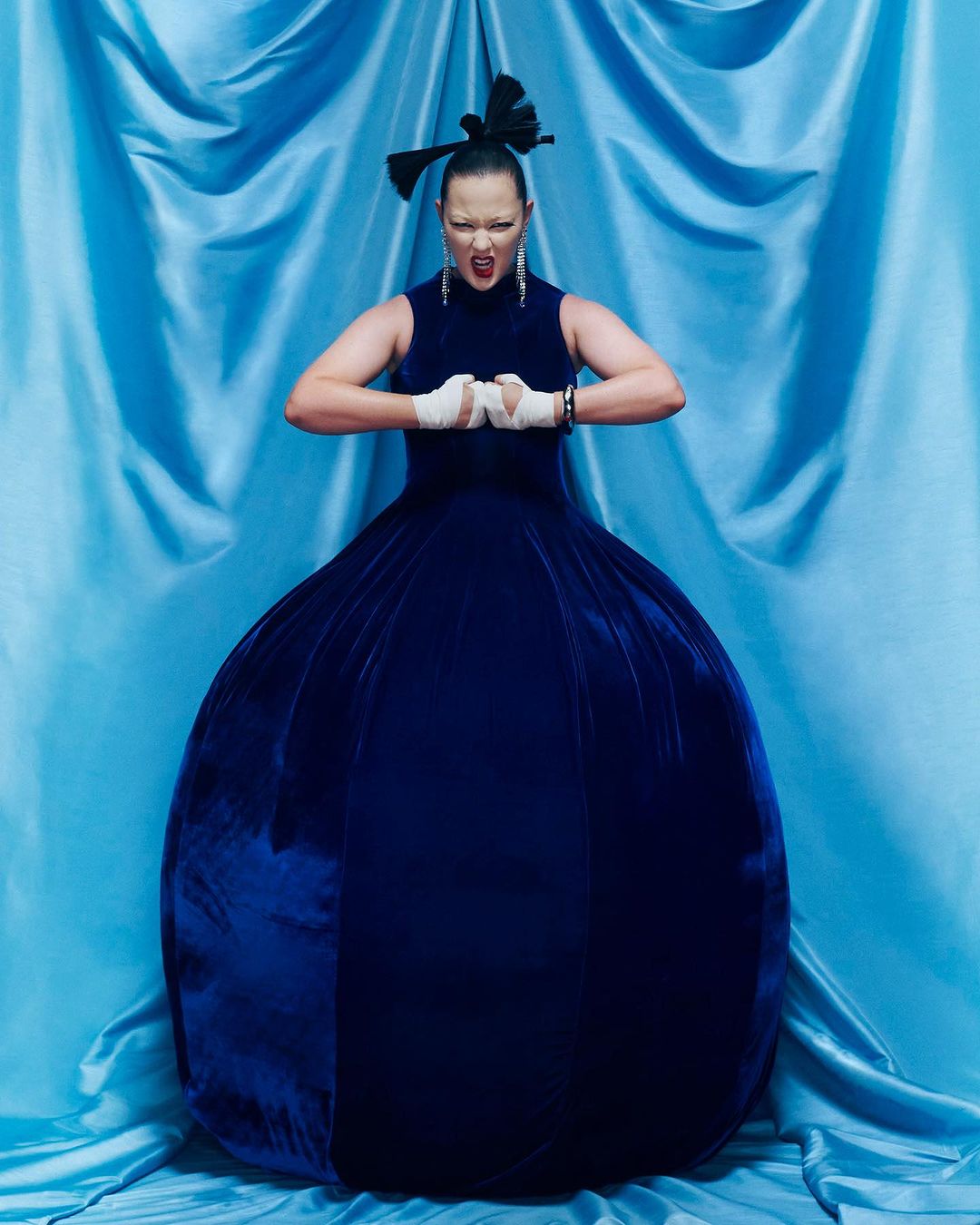 A woman with clenched fists, wrapped in white bandages, wears a blue velvet dress against a draped turquoise fabric backdrop.