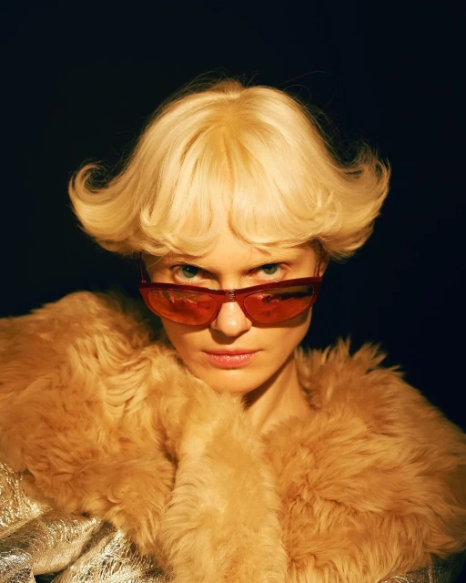 Close-up portrait of a blonde woman wearing red sunglasses and a tan faux fur coat against a dark background.