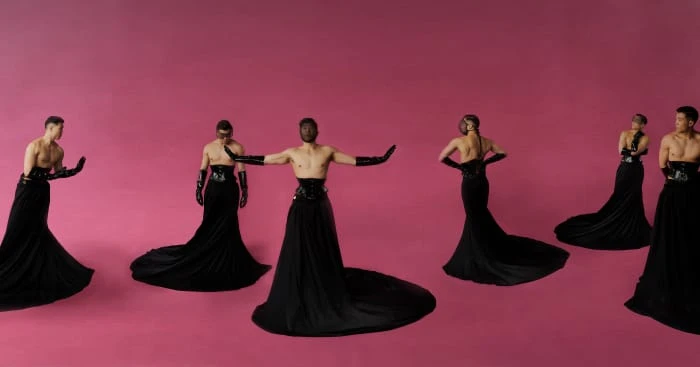 Multiple shirtless male fashion models wearing black dresses and gloves on a pink background, showcasing fashion and performance art.