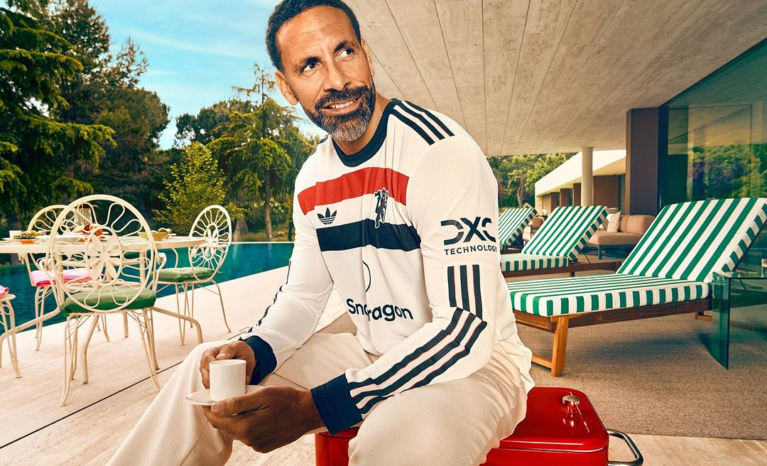 Rio Ferdinand sits outdoors on red stool, holding teacup in fashion shot with architecture, trees, pool, and lounge chairs.