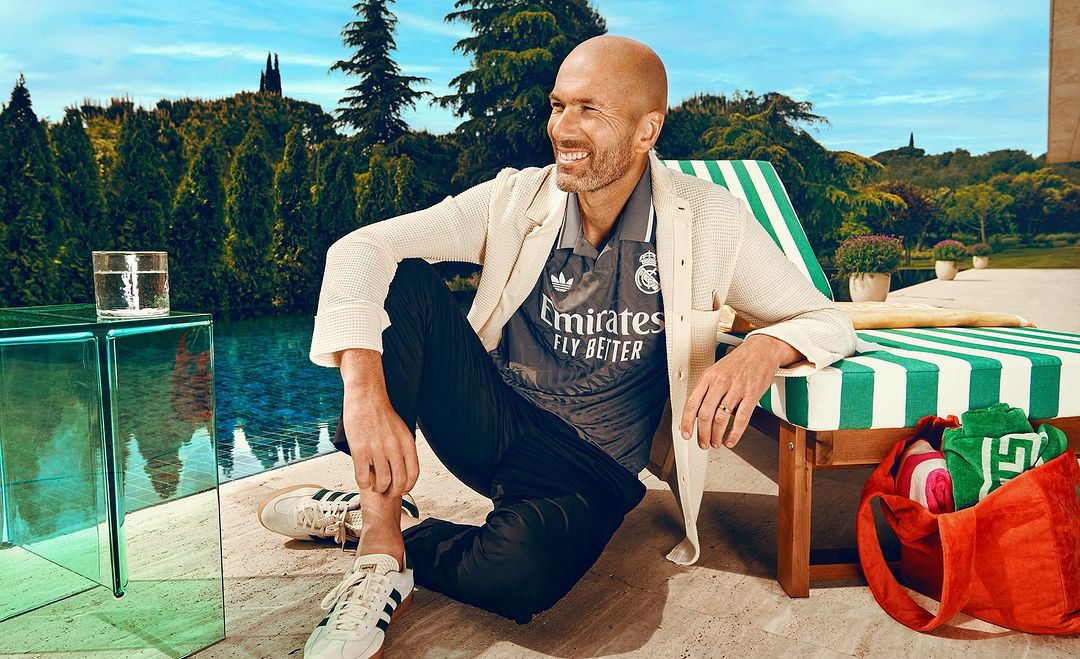 Man in soccer jersey poses outdoors by pool with trees and blue sky. He is seated next to a striped lounge chair and glass table.