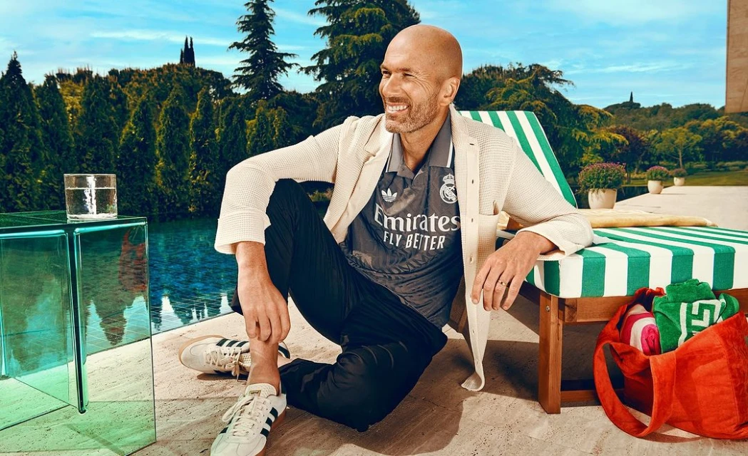 Man in soccer jersey poses outdoors by pool with trees and blue sky. He is seated next to a striped lounge chair and glass table.