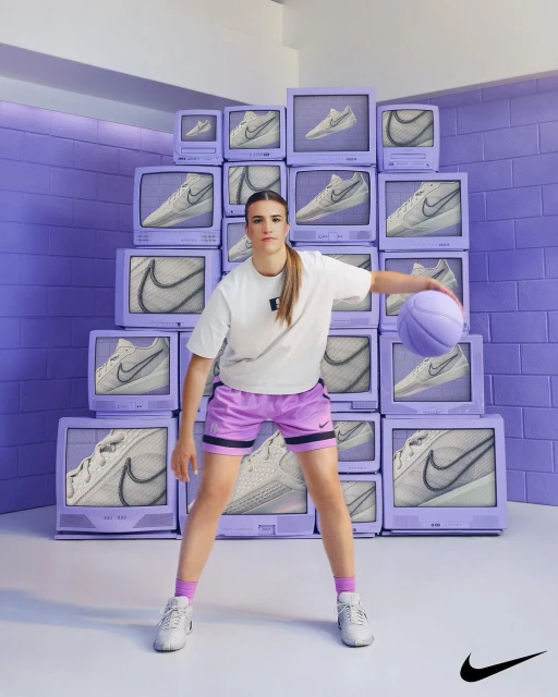 Female basketball player with ball stands in front of stack of purple televisions displaying images of Nike sneaker; indoor studio shot.