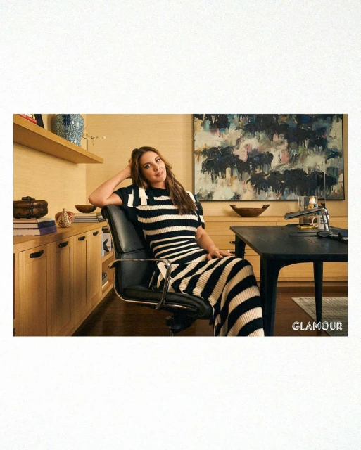 A woman sits in an office chair, wearing a black-and-white striped dress, beside a desk. Visible in the background are shelves and art.
