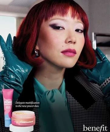 Close-up of an Asian woman with red hair wearing makeup, teal gloves and shirt, and a black and white jacket, promoting Benefit cosmetics.