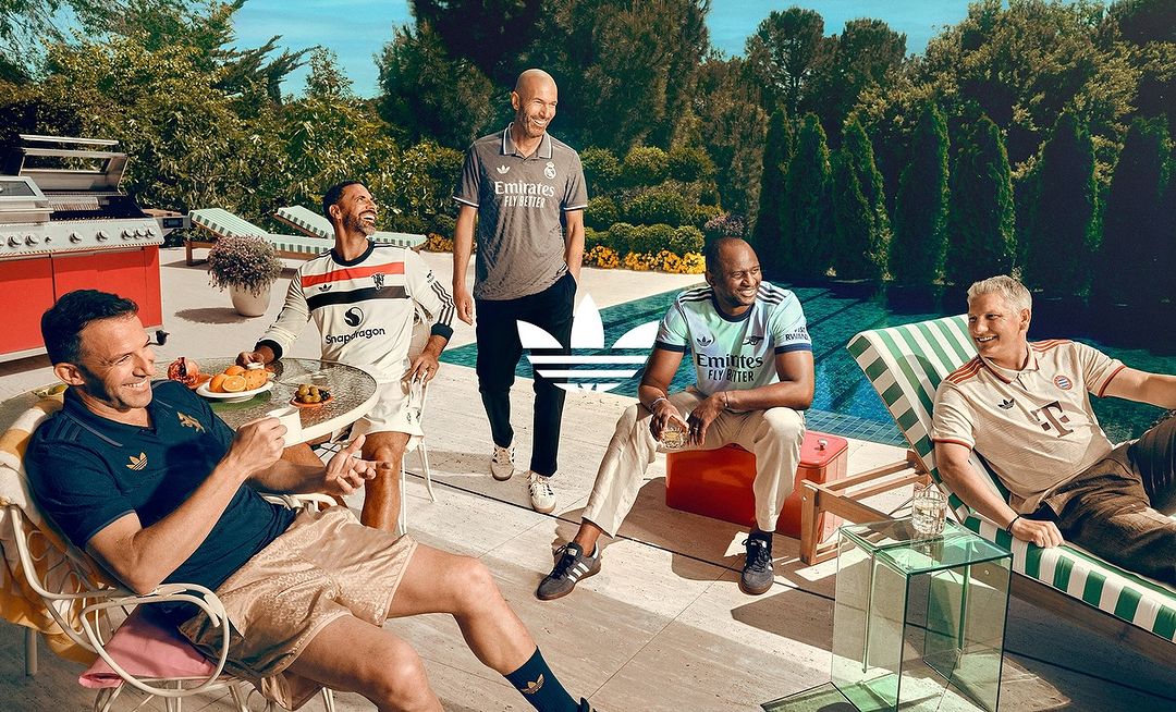Group of soccer legends relaxing poolside in Adidas apparel, featuring Zidane, with lush greenery, bright lighting, and brand visibility.