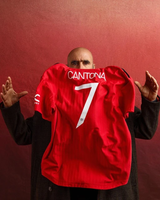 Bald man in dark coat holds up red Manchester United jersey with name Cantona and number 7 against a textured red backdrop.