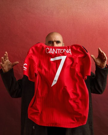 Bald man in dark coat holds up red Manchester United jersey with name Cantona and number 7 against a textured red backdrop.
