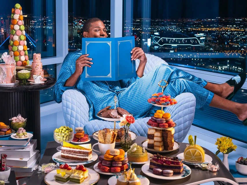 Adult male in pajamas lounges in a blue chair reading a book, surrounded by an abundance of colorful cakes and desserts at night.