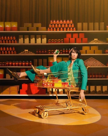 Person in green suit reclines in a gold shopping cart in a supermarket aisle against shelves stocked with orange and brown objects.