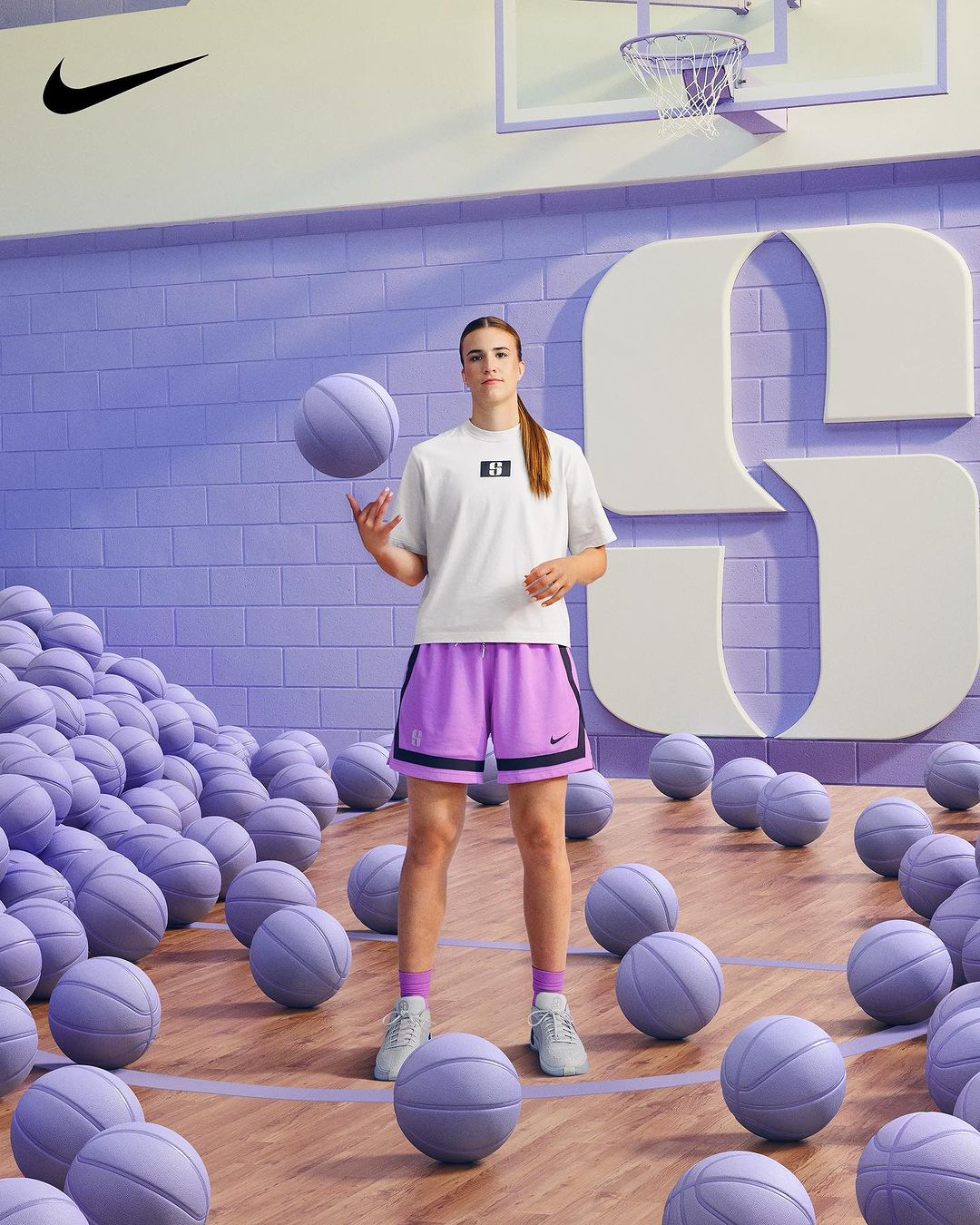 Female athlete in athletic wear stands in a basketball court surrounded by basketballs, tossing one in the air, Nike logo in view.