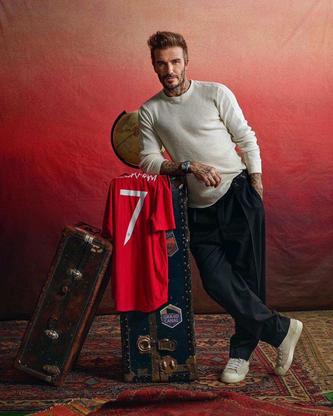 David Beckham poses next to vintage luggage with a red #7 soccer jersey and a globe in a studio environment with dramatic lighting.