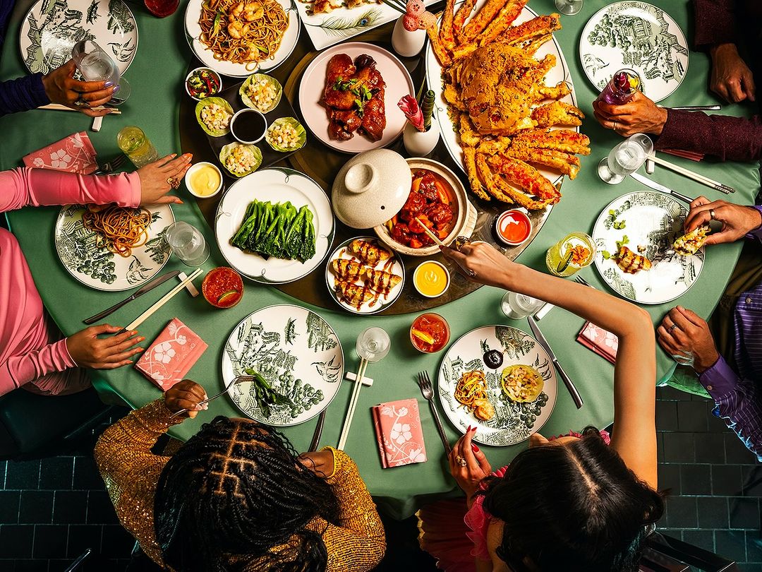 Overhead view of a round table laden with various Asian dishes, decorative plates, cocktails, chopsticks, and several seated diners.