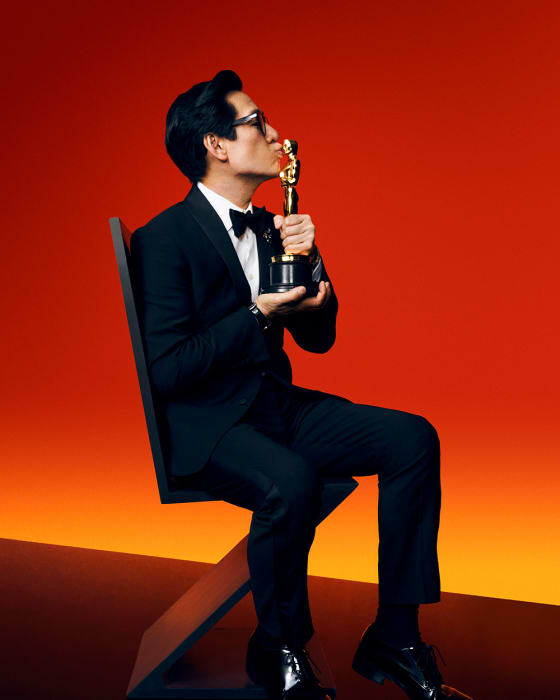 Adult male in a black suit seated kissing an Oscar statue in a photo studio with gradient orange and black background. Black chair visible.