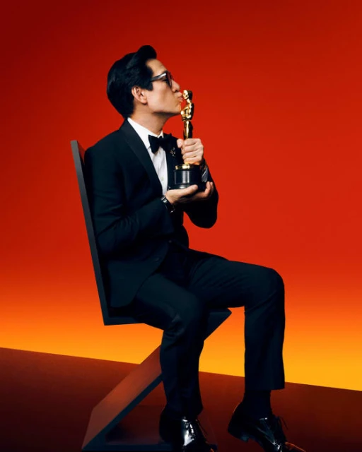 Adult male in a black suit seated kissing an Oscar statue in a photo studio with gradient orange and black background. Black chair visible.